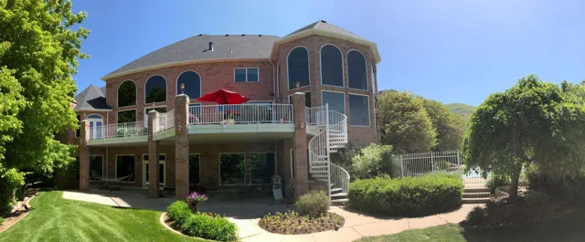 Large brick house with a deck, spiral staircase, and green lawn under a blue sky.