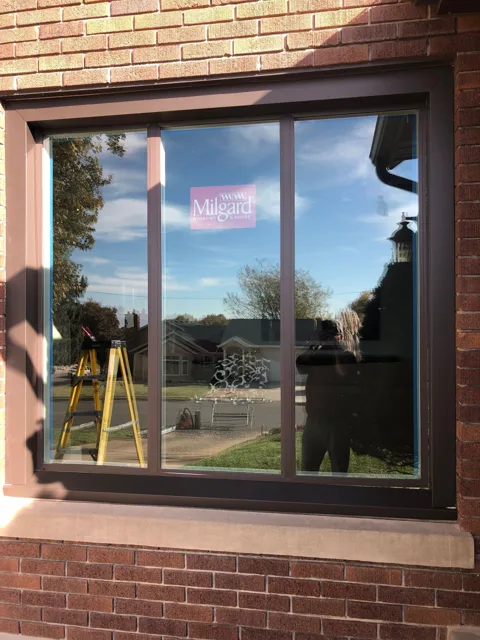 Large, brown-framed window reflecting sky and yard. A red window sticker is visible. Brick building exterior.
